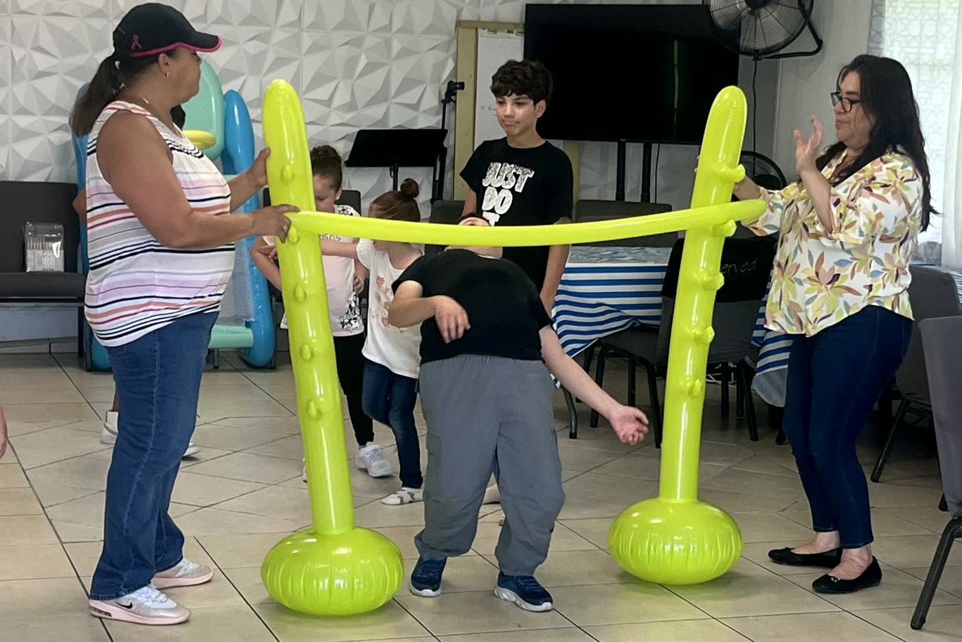 Dos mujeres de la iglesia sostienen un arco de globos mientras los niños pasan por debajo, en el salón de compañerismo
